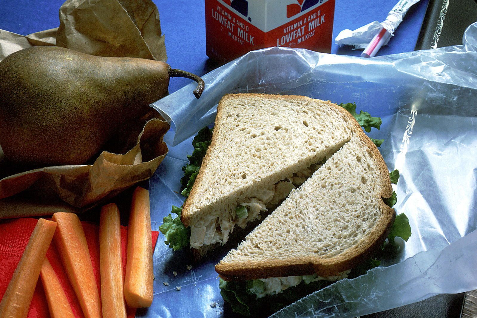 "A lunch sits on a blue tablecloth with a brown paper bag and red napkin. There are carrots, a pear, a sandwich on wheat bread with lettuce (chicken salad) and a carton of milk." Image courtesy the National Cancer Institute, 1989, via Wikipedia.